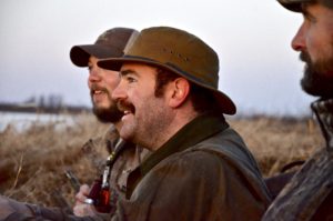 Two men in a field with hats and drinking.