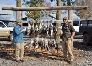 Two men standing next to a rack of dead birds.