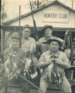A group of men holding dead birds in front of a hunter club.
